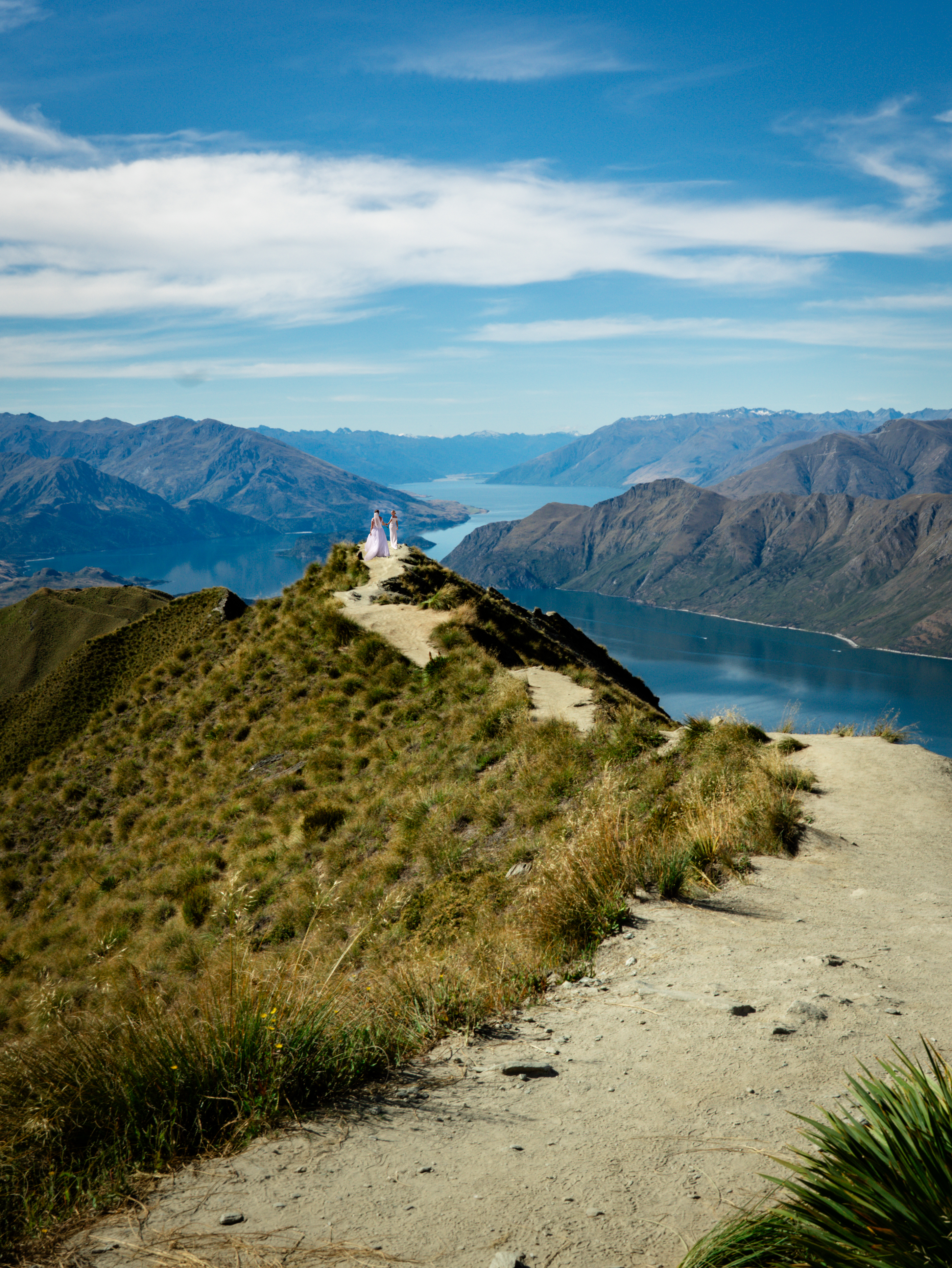 Queer couple walking at the edge of a cliff with an overview of lakes and mountains in New Zealand.